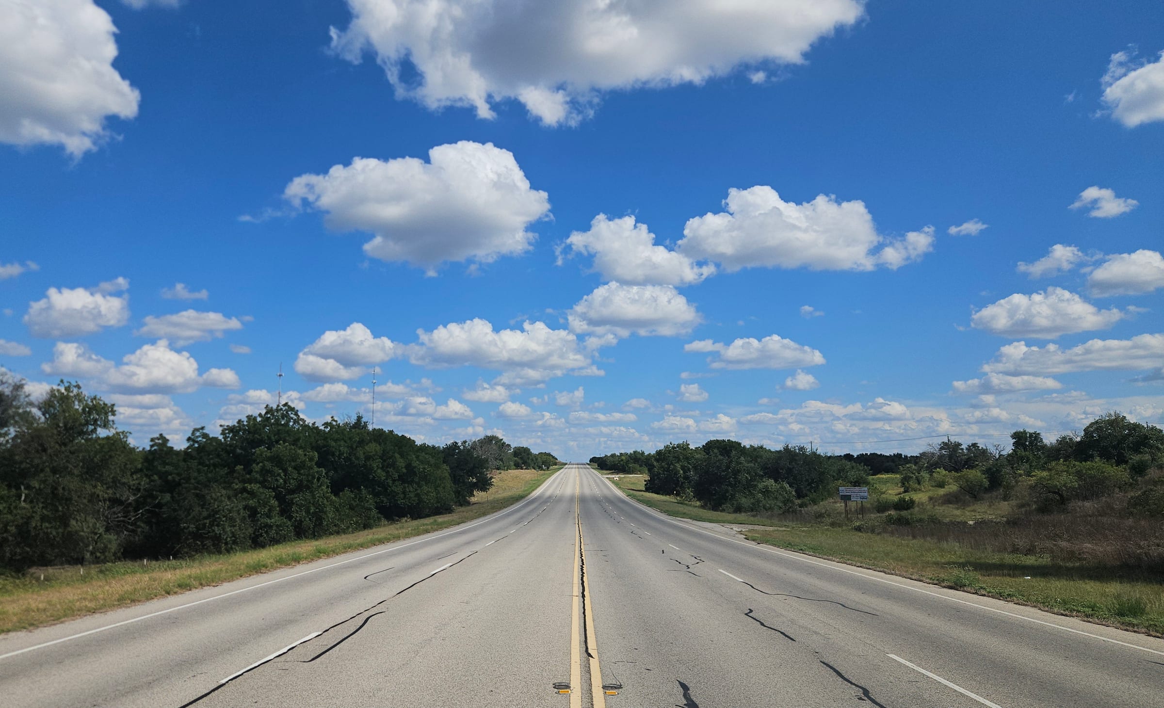 An empty highway running straight to the horizon between green trees, under a bright blue sky full of small white clouds